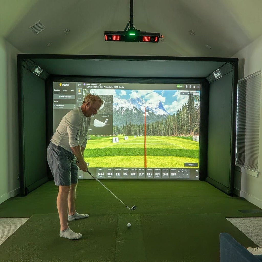 Person playing golf in a virtual reality simulator with a large screen displaying a golf course.